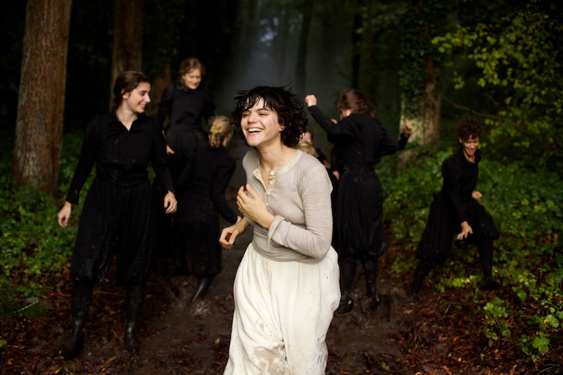 A group of women in black smile and run through the forest. A woman in white leads them.
